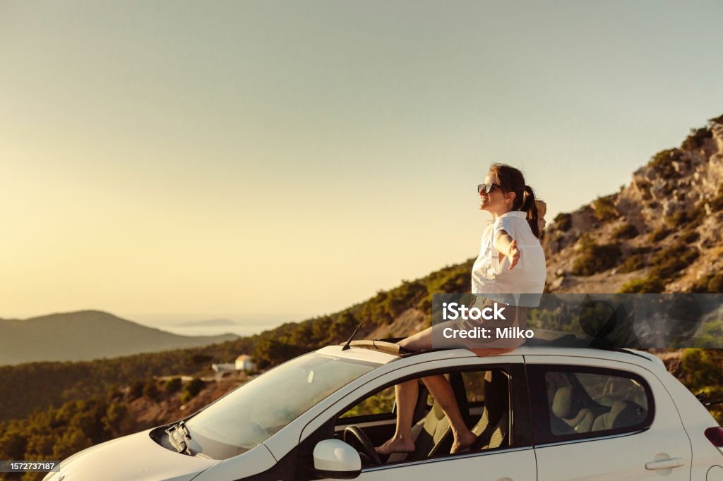 Woman enjoying the beautiful sunset o na road trip Shot of a woman enjoying a relaxing road trip. She is taking a break from a car drive and enjoying the sunset Car Stock Photo
