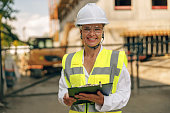 Female engineer in protective helmet making notes on construction site and looking at camera