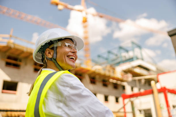 Smiling female construction worker in protective helmet standing against on construction background stock photo