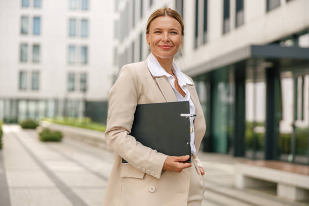 istock Female manager is standing on office building background with documents and looking at camera 1572768032