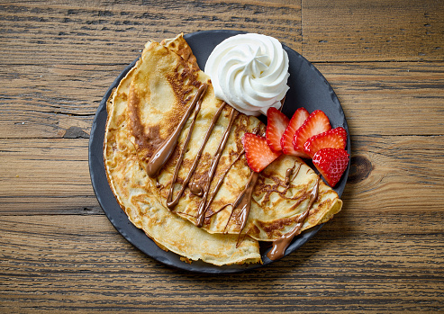 freshly baked crepes with whipped cream and strawberries on wooden table, top view