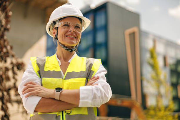 Smiling female construction worker in protective helmet standing against on construction background stock photo
