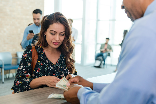 The mid adult woman watches and listens to the unrecognizable bank teller count her money that she just withdrew from her account.