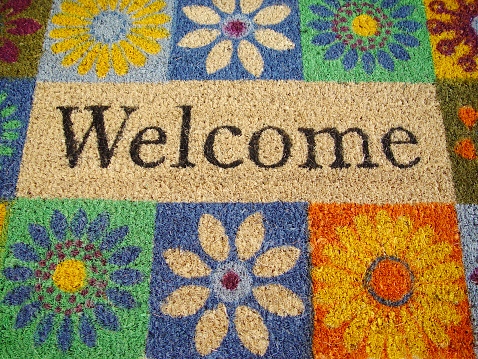 WELCOME written on a bight colored and with flowers decorated doormat.  Images taken from above with the word welcome in the center. The floral pettern is multi colored.
