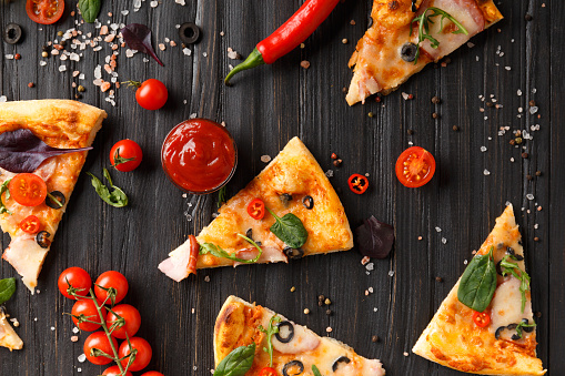 Pizza slices with jerky, olives, cheese and herbs are laid out on a dark wooden background, a branch of fresh cherry tomatoes, sauces and seasonings for pizza top view. Italian food.