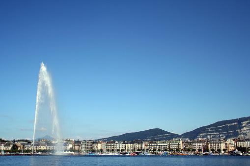 view of jet d'eau fountain from across Lake Geneva on a clear sunny day.  City buildings are visible on the lakeshore