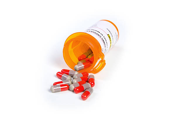 An open prescription medicine bottle pouring out capsule style pills on a white background.