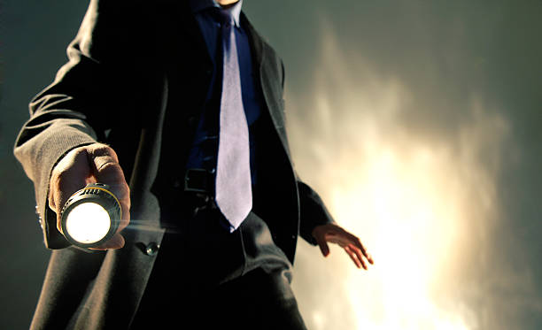 Man in Suit Holding Torch A close up of the torso of a Caucasian man dressed in a dark business suit, shirt and tie holding a battery operated torch pointed towards the camera, with his arm out in a dramatic pose. The man is standing against dark background with a brightly lit abstract pattern.  searchlight photos stock pictures, royalty-free photos & images