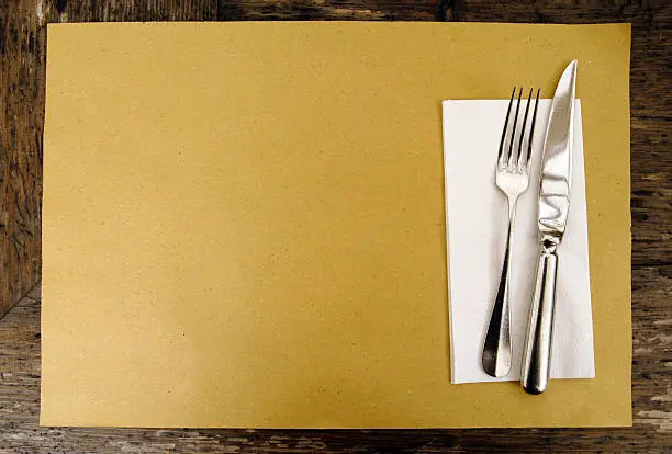 A casual place setting consisting of knife and fork, white napkin and a brown paper place mat.