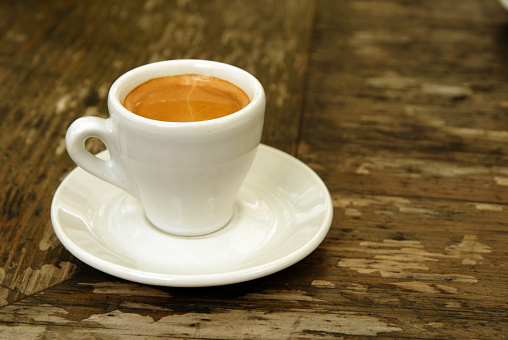 A white cup with espresso coffee positioned left of frame on a wooden table.