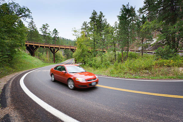 Red Sedan Car Driving Scenic Mountain Highway with Pigtail Bridge A red sedan driving a scenic mountain highway towards a pigtail bridge located near Mount Rushmore in the Black Hills, South Dakota, USA. The National Memorial and Parks are favorite summer road trip travel destinations for American family tourists. Visitors can explore forests and enjoy winding roads with hairpin curves. driving winding road stock pictures, royalty-free photos & images