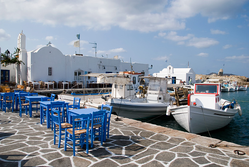 Blue tables and chairs of a quay side restaurant at the harbor of Naoussa in Greece. A very traditional greek scene with fishing boats an a small white church. Naoussa is a small town on Paros, one of the Cyclades Islands in Greece.