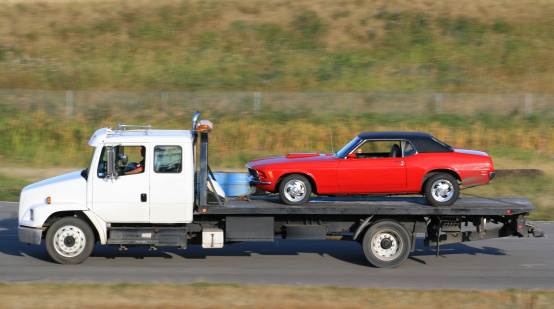 A red sports car being towed by a tow truck. Hauling, freight, or trucking theme. This image is a pan shot. Flat deck truck with automobile. Rural highway. White heavy duty truck with winch and loader. Flatbed. 