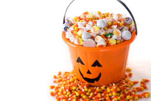 Pumpkin bucket overflowed with Halloween candy. Candy corn is spread all around. On white, copy space.