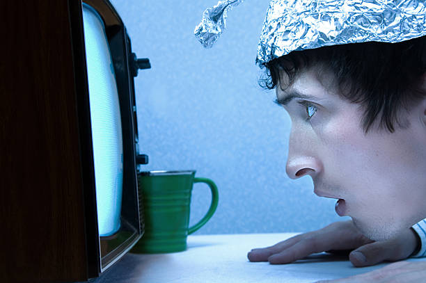 Paranoid Slightly. A young man wearing a tinfoil hat and watching a fuzzy old TV set. Probably seeing aliens. tin foil hat stock pictures, royalty-free photos & images