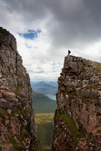 A climber on the edge of a rocky precipice with lochs and mountains behind