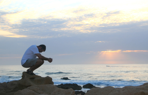 A young Caucasian man prays beside the sea. Los Cabos, Mexico. Side view of handsome white man squatting and meditating beside the sea. Additional themes in the image include spirituality, balance, salvation, Christianity, faith, religion, hope, gratitude, prayer, thinking, contemplation, folding hands, morning, misty, purity, single, trouble, depression, self, vitality, wellness, and health. 