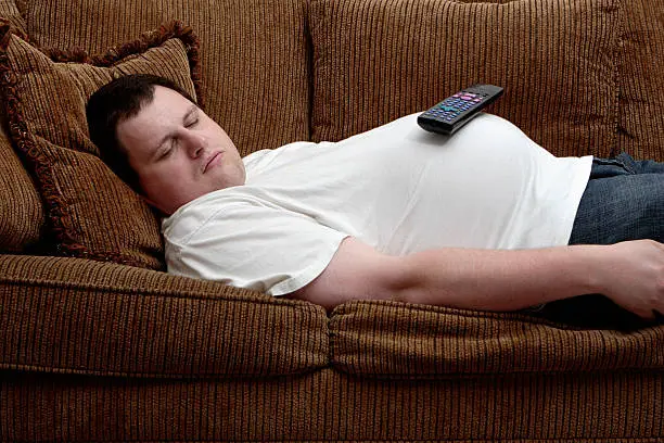 A man sleeps on the couch, with a TV remote control balancing on his protruding stomach.