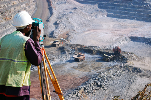 An African man, locally employed as a surveyor at an open-pit copper mine in Zambia, peers through his survey instrument.  This work records the daily changes in the open-pit, and help guide mining activities to the engineer's plans.