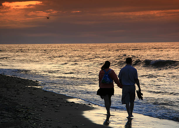 Couple on a Romantic Beach Walk A couple on a romantic walk on the beach. A silhouette of a middle aged Caucasian couple walking on a beach with a beautiful sunset. Image location is PEI, Canada on Cavendish Beach. Man is holding sandals and they are both wearing shorts on a warm summer evening. Back view. Models are unrecognizable. Two people in the image.  cavendish beach stock pictures, royalty-free photos & images