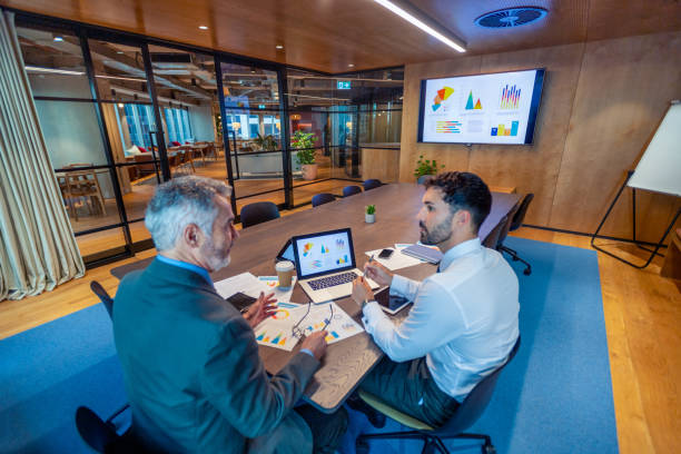 istock Two business men meeting in a board room. There is technology and documents on the conference table including a laptop 1572935855