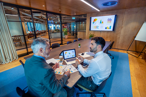 Two business men meeting in a board room. There is technology and documents on the conference table including a laptop. There is a t.v. with finance presentation. Documents show finance data charts and graphs.