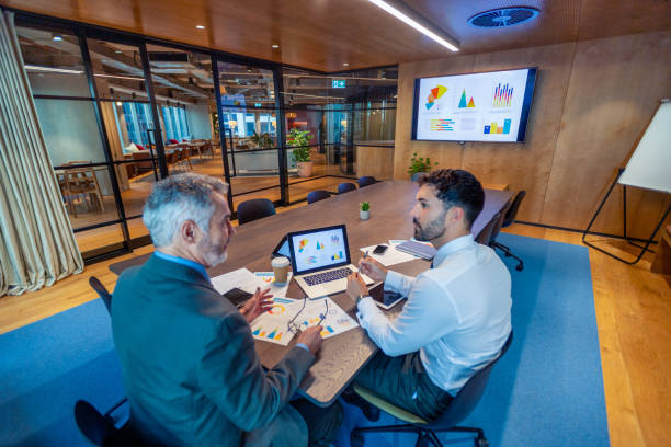 Two business men meeting in a board room. There is technology and documents on the conference table including a laptop stock photo