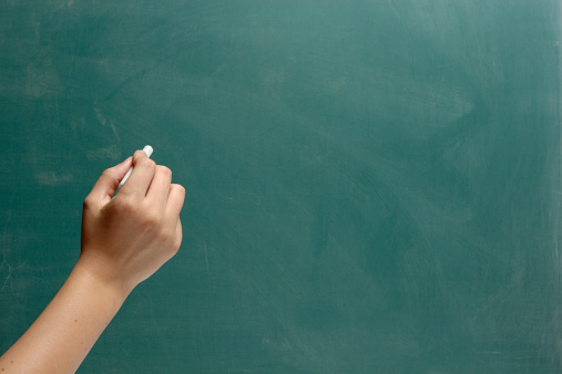 A female hand holding chalk, moves to write something on a blank blackboard.