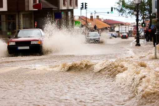 A road in Bansko, Bulgaria, awash with water during a flash flood after heavy storms.  Car driving through flood water as river bursts its banks during a summer storm.  Waves are produced by the rushing water and spray is produced by the car wheels as it drives towards the camera. Taken in Summer.