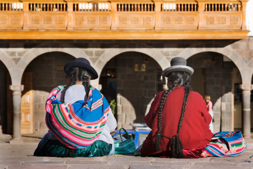 Two Peruvian women sitting on a town square plaza ledge in Cuzco, Peru. Their backs are turned and they are dressed in a traditional manner, wearing braided long hair and wool hats and wrapped in colorful blankets and sweaters.