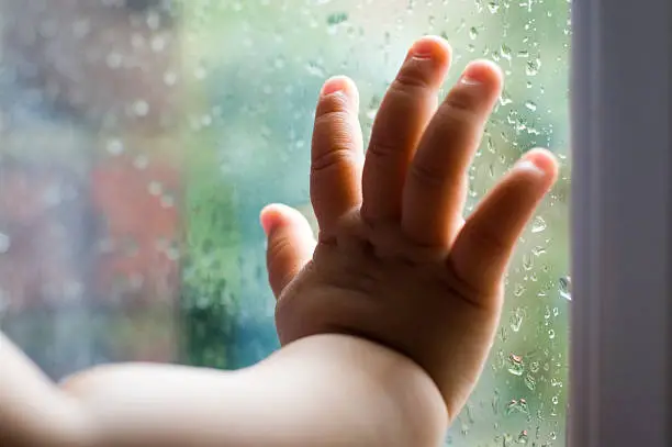 Baby is touching window with her hand. It’s a rainy day hence the droplets of water on the outside