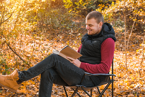Young man is sitting on chair in an autumn park or forest and reading book. Mental recreation in nature. Quiet country life. Escapism. Digital detoxification. Concept of reading book in an autumn park