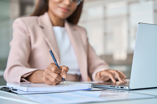 Close up view of professional busy African American business woman manager executive working on laptop computer writing notes in notebook at work elearning in corporate office.