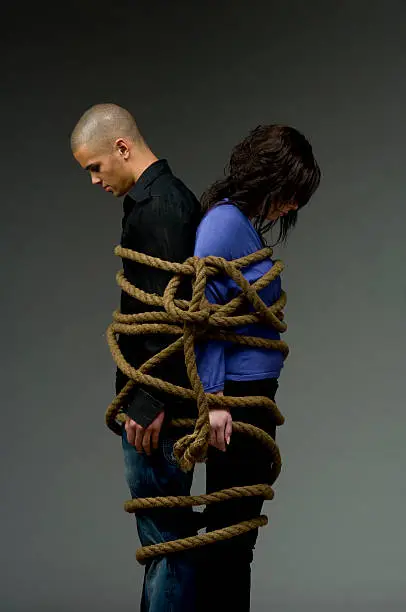 Dark haired young woman and a man with a shaved head tied together by a hawser with a big knot looking very down as they are fighting