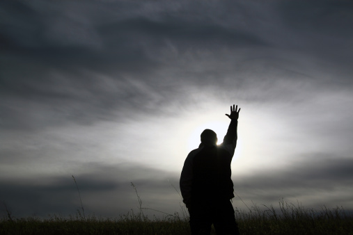 A man lifts his hand to heaven. Black and white silhouette of many in praise and worship position. Hand lifted to heaven towards god. Unrecognizable man in his 30s. Additional themes include forgiveness, Christianity, balance, spirituality, prayer, meditation, redemption, salvation, easter, godliness, righteousness, hope, healing, and faith. 