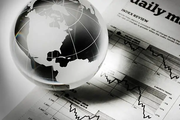 A glass globe paperweight resting on newspaper financial page charts and graphs, which indicate downward global business, investment, and finance trends. The earth sphere casts a shadow, implying recession and forecasting stock market trouble. The USA, Canada, parts of Europe and the Americas are visible, for concepts of world economy.