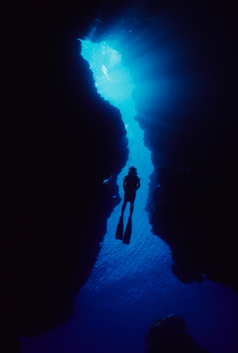 Silhouetted free diver swimming through school of fish in underwater cave into bright light