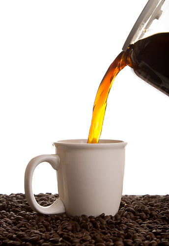 Coffee being poured into a coffee cup sitting on coffee beans against a white background.