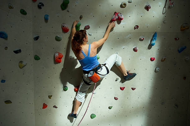 Female climber purposeful climbs up the indoor climbing wall. The climber has good physical training and relief muscles. She has insurance. The athlete is dressed in a red top and black short shorts.