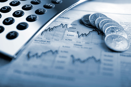 A close up, blue tinted conceptual image of silver calculator and British ten pence coins on top of a generic financial newspaper page showing stock market graphs. The image illustrates the concepts of: finance, money, economy, stocks and investment. Shot with a shallow depth of field. 