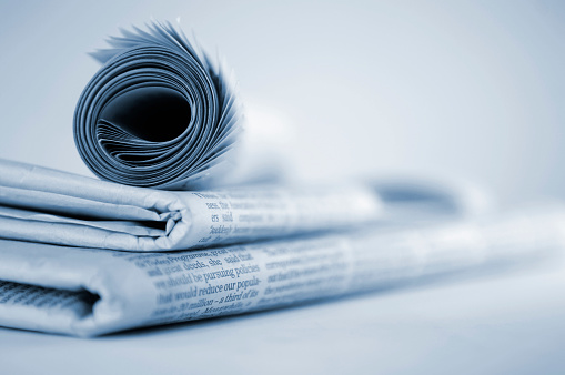 A blue tinted image of a pile of newspapers stacked flat on a flat surface with a rolled up newspaper on top. Shot with a very shallow depth of field, with focus on the left side of the image. The newspapers are against a plain background. With copyspace. 
