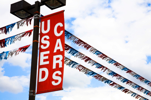 'Used Cars' sign over a vehicle dealership car lot. Red. Clouds in sky. 