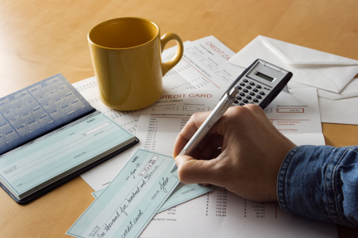 Writing checks and paying bills for credit card debt. A human hand holds a pen to the check blank. A nearby calculator and stacks of statements suggest calculation of home finances and cash flow management for addressing problems and taking action for responsible solutions in personal spending.