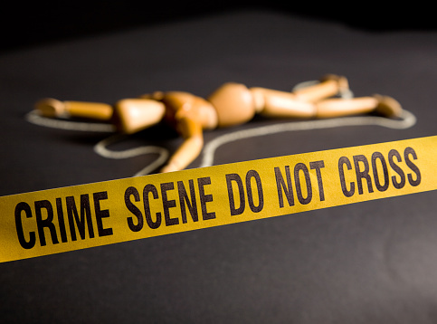 A wooden artist's figure laying in a chalk outline behind crime scene tape.  Harsh directional lighting.