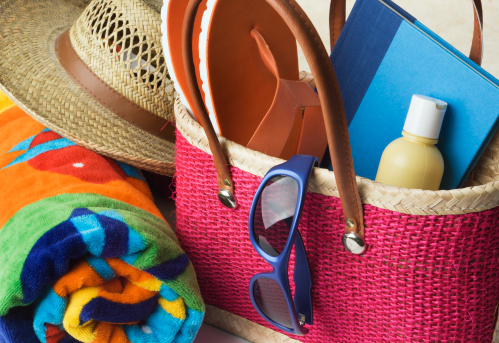 Summer vacation beach bag loaded with supplies. The rattan purse container holds suntan lotion, moisturizer or sunscreen, sunglasses, towel, flip flops, a good book or trashy novel, and straw sun hat, in preparation for ocean recreation, fun in the sun, and getting away from it all. Close-up with no people.