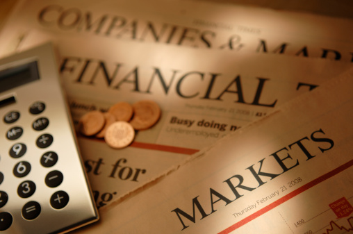 A close up image of a silver calculator, and some British sterling penny coins resting resting on loosely arranged sections of a financial broadsheet newspaper.  Shot with a shallow depth of field with focus on the foreground ‘Markets’ section of the newspaper. 