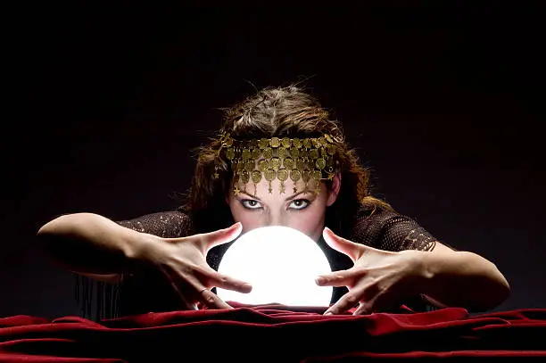 Mysterious focused fortune telling woman wearing a copper hair dress with her hands on a glowing crystal ball looking at the camera