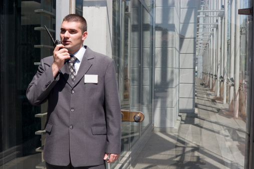 security guard standing at the glass door of corporate building talking with walkie talkie in his hand