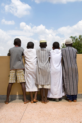 Rear view of five young african boys standing next to each other. The boys are hanging and looking over a wall. Four are dressed in traditional muslim dresses and one in eastern clothing.