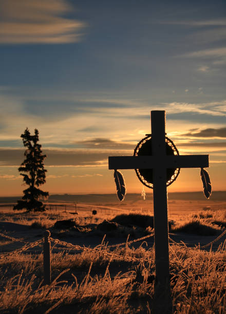 A large stone cross monument rises against a blue sky filled with clouds, bright sunlight, and floating particles or bokeh effects creating an ethereal atmosphere. This image symbolizes faith, hope, and spiritual transcendence, representing the connection between earth and heaven, memorial remembrance, and divine presence.